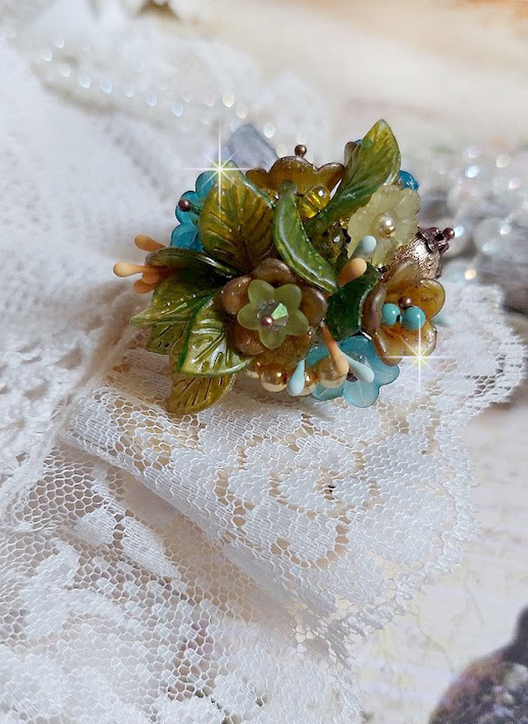 Bague Cézanne montée avec des petites fleurs lucites peintes à la main à laquelle s’ajoute des feuilles, cristaux, perles, étamines, grosses rocailles, perles nacrées et métal sur une bague en Laiton