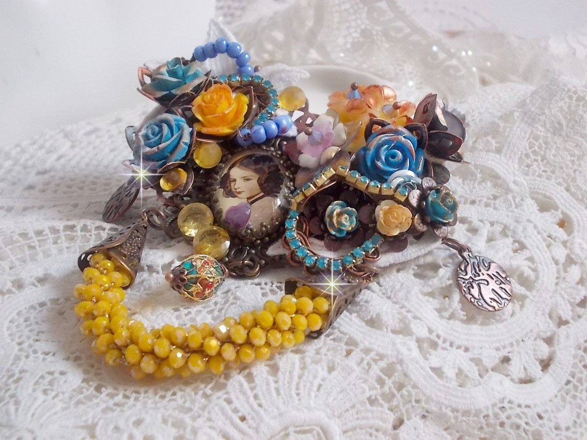 Broche Eclats Pétillants d’inspiration Baroque, ornée de fleurs en résine autour d’un cabochon strassés et perlés sur estampes, cristaux, breloques, perle cloisonnée, perles jaunes en verre et roses. 
