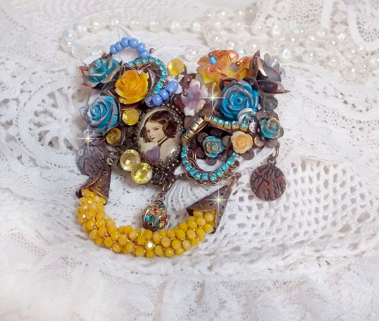 Broche Eclats Pétillants d’inspiration Baroque, ornée de fleurs en résine autour d’un cabochon strassés et perlés sur estampes, cristaux, breloques, perle cloisonnée, perles jaunes en verre et roses. 