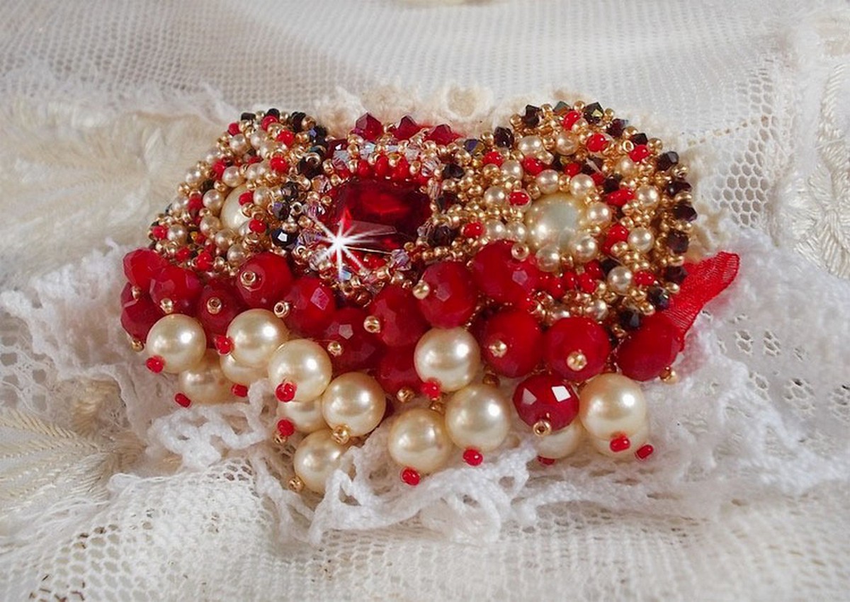 Broche Rubis brodée avec trois cabochons Ivoire et Rouge à laquelle s’ajoute des toupies aux couleurs Cristal, Burgundy et Garnet, des perles nacrées et opaque. Monté sur une dentelle et une broche