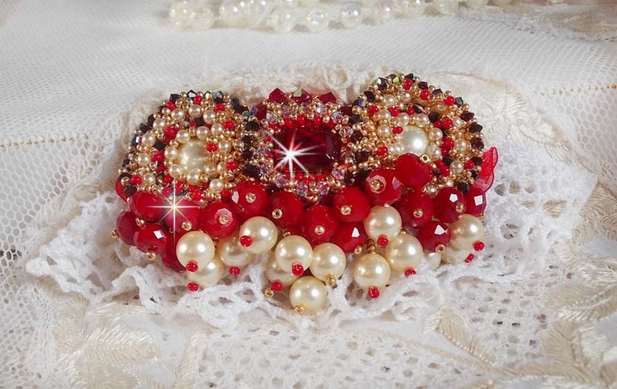 Broche Rubis brodée avec trois cabochons Ivoire et Rouge à laquelle s’ajoute des toupies aux couleurs Cristal, Burgundy et Garnet, des perles nacrées et opaque. Monté sur une dentelle et une broche